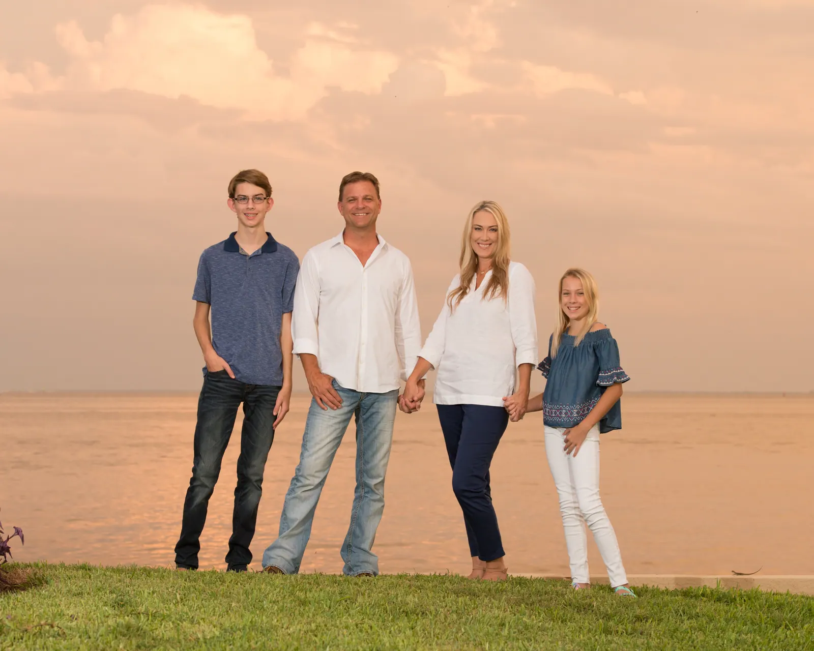 Family of four standing on a waterfront lawn at sunset — family portrait by Moorman Photographics
